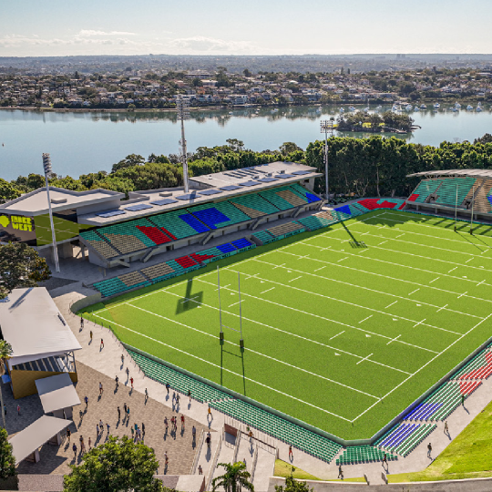 Ariel shot of a large sporting oval with bright green grass framed by trees and ocean. People walk on paved areas around the oval